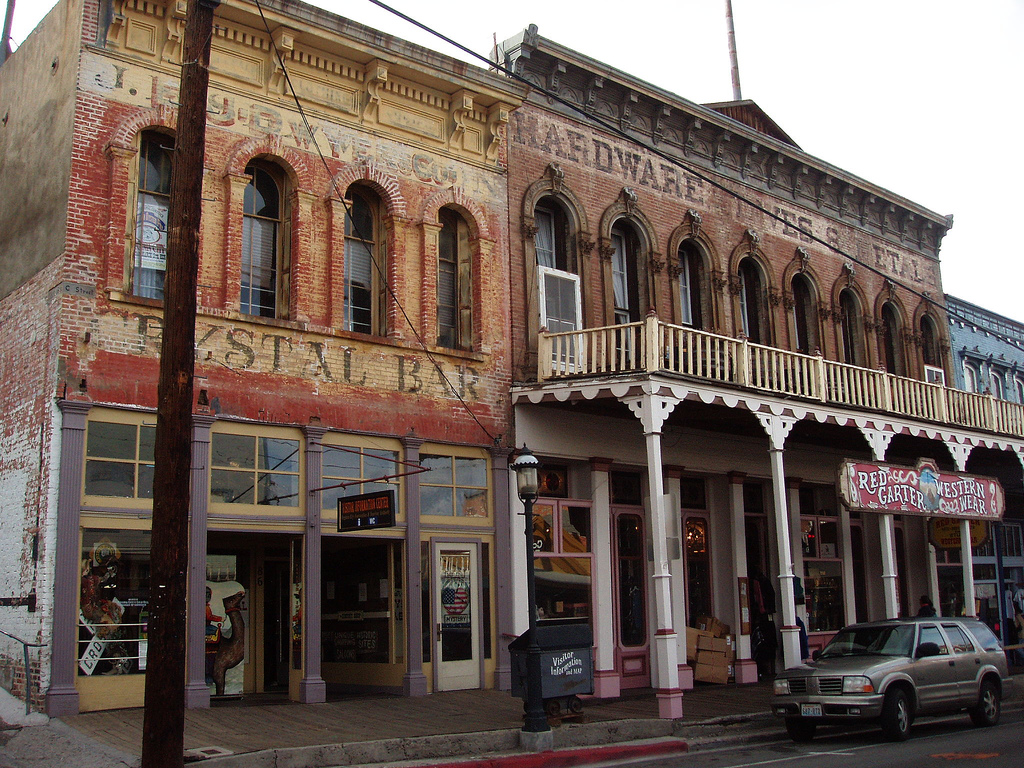 Historic brick buildings with faded painted signs and arched windows line a street with a covered wooden sidewalk and a parked car in front.