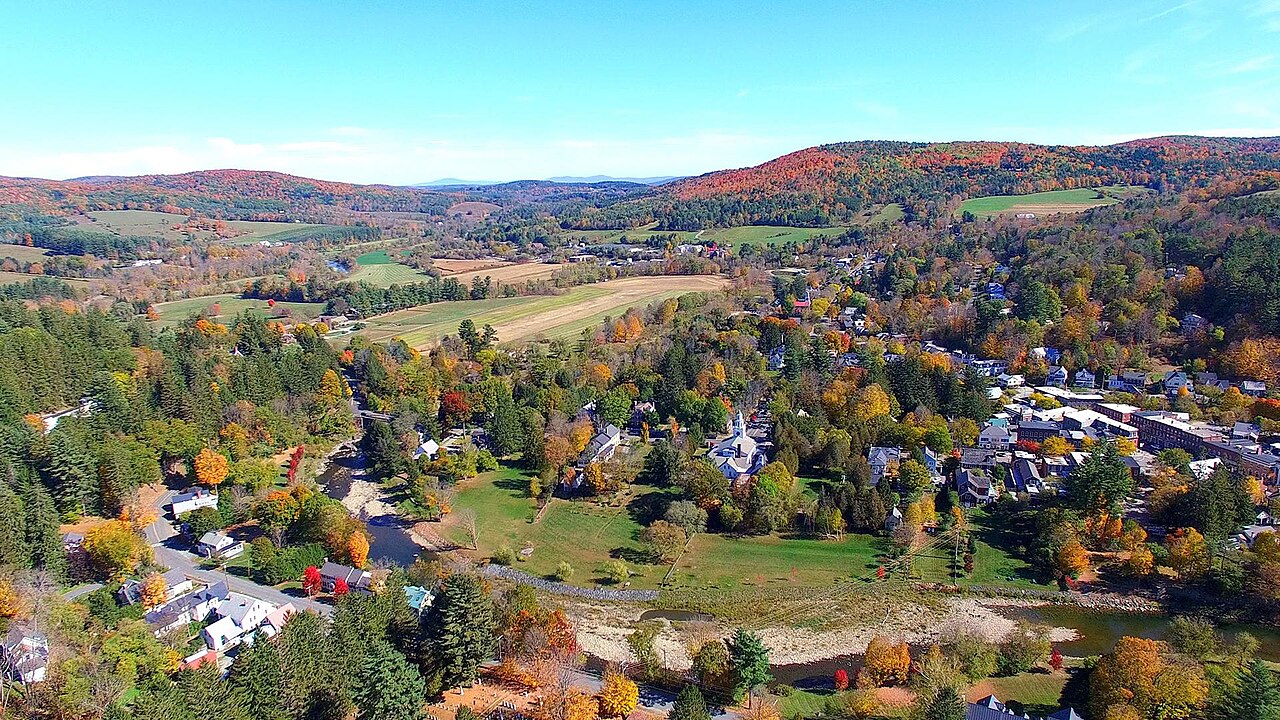 Aerial view of a rural town amid autumn trees, fields, and rolling hills beneath a clear blue sky.
