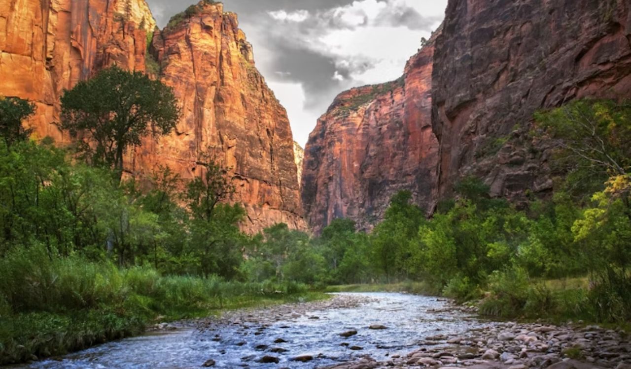 A river winds through a green valley bordered by tall red cliffs beneath a partly cloudy sky.