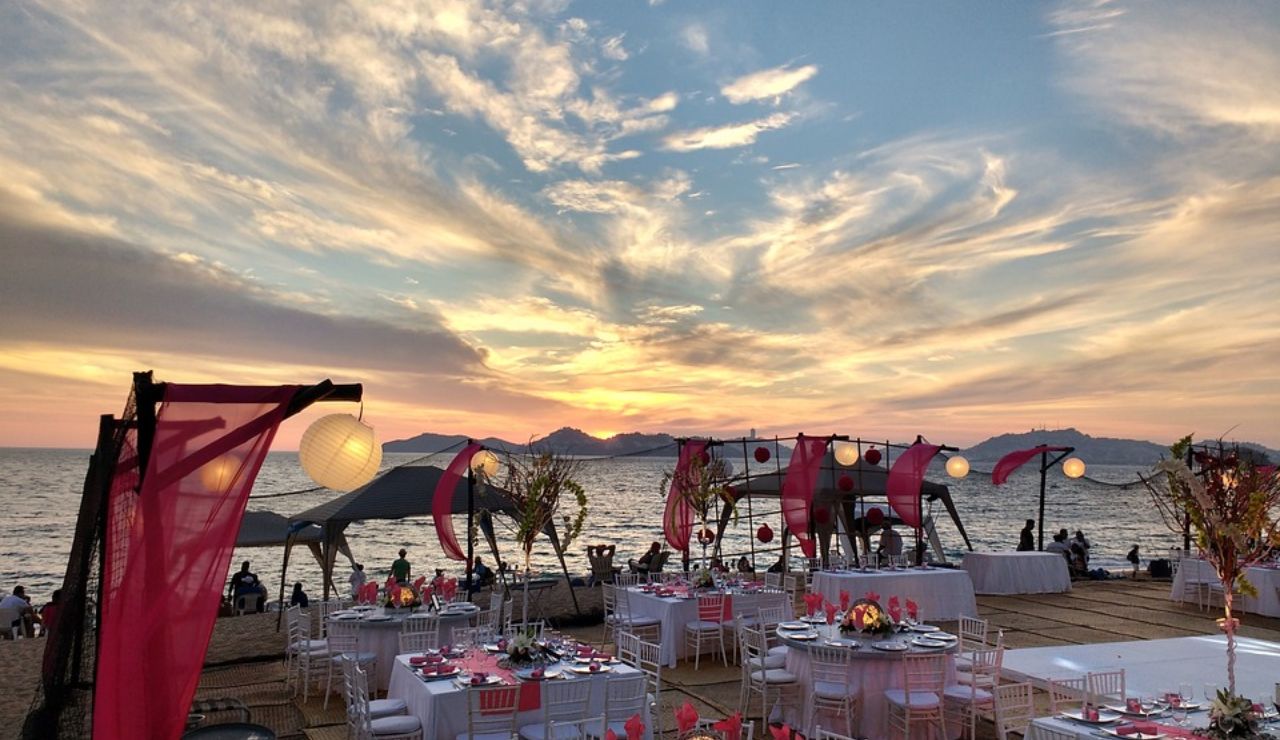 Romantic beachside wedding reception setup at sunset in Acapulco, Mexico, with decorated tables, pink drapes, and ocean view