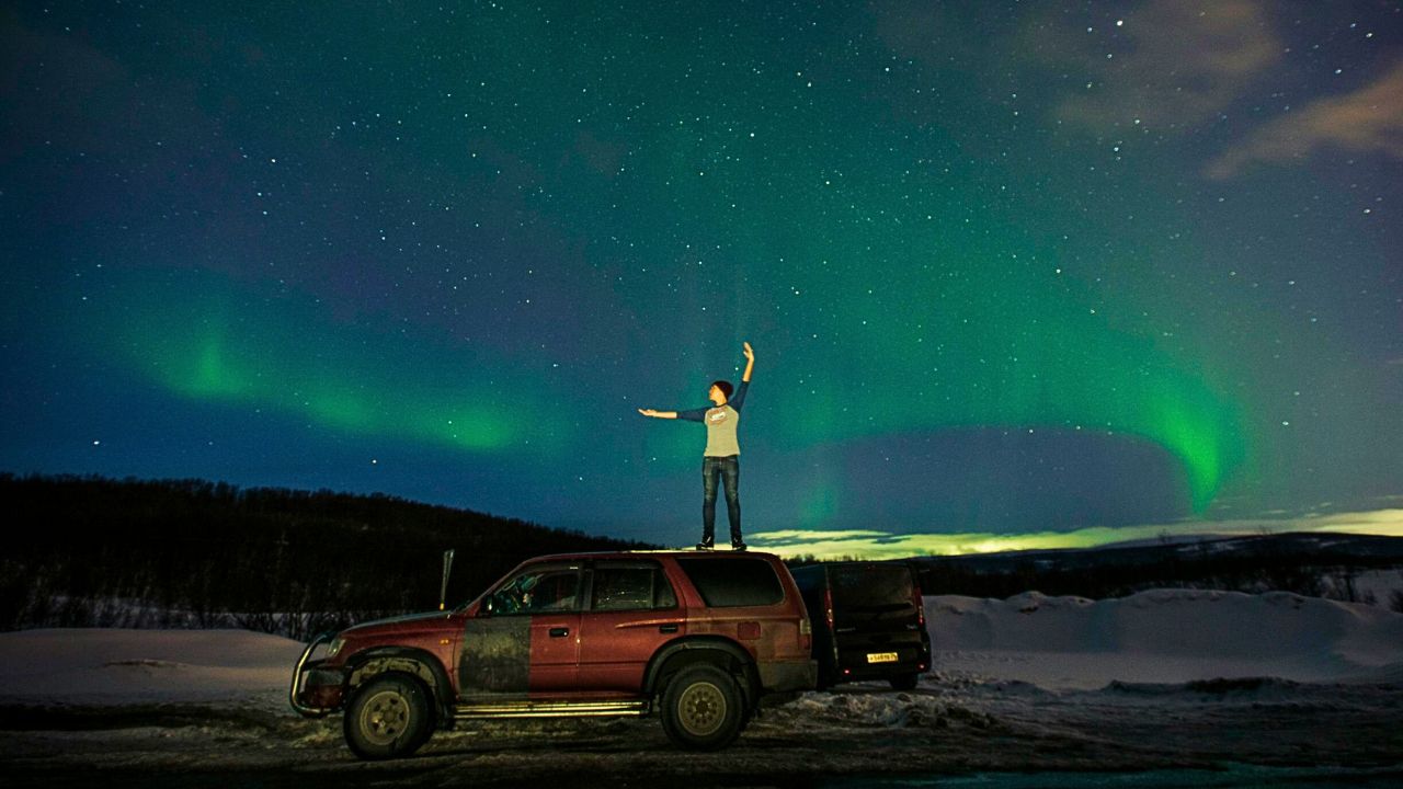 A traveler stands on a car in Alaska, overlooking a vast glacier valley surrounded by snowcapped peaks under a wide sky.