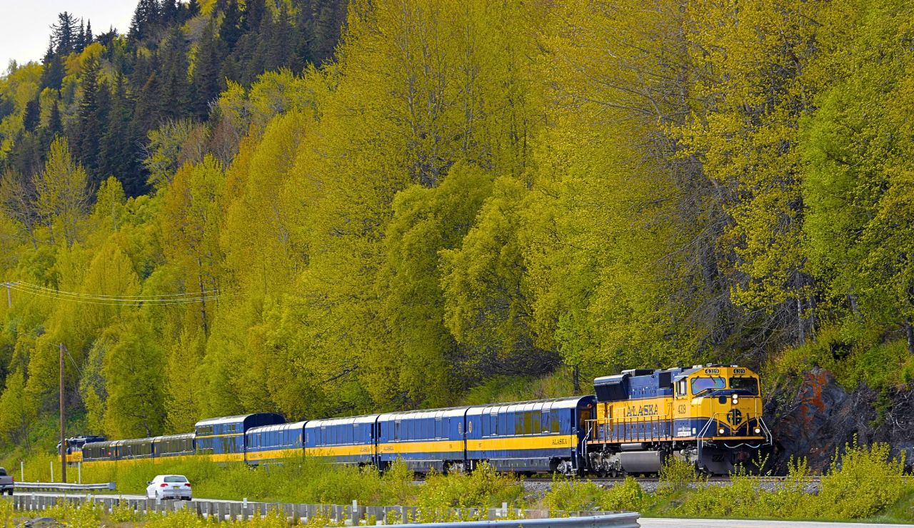A yellow and blue Alaska Railroad train travels through mountain scenery with panoramic windows reflecting the landscape.