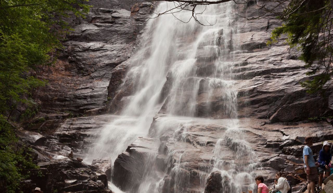 A wide, graceful waterfall flows down tiered, dark rock formations, while a few people rest nearby, soaking in the tranquil, misty scene.