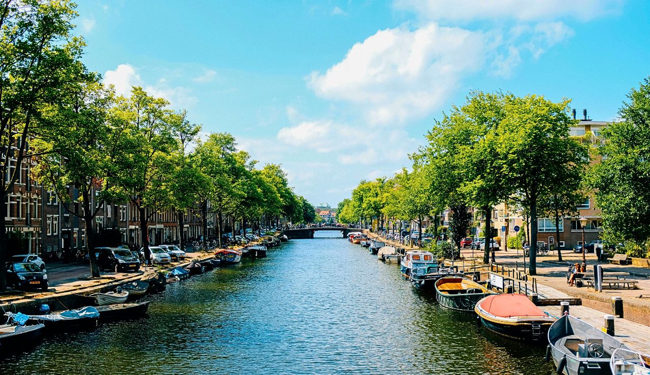 Boats line a canal bordered by trees, parked cars, and buildings beneath a partly cloudy sky.
