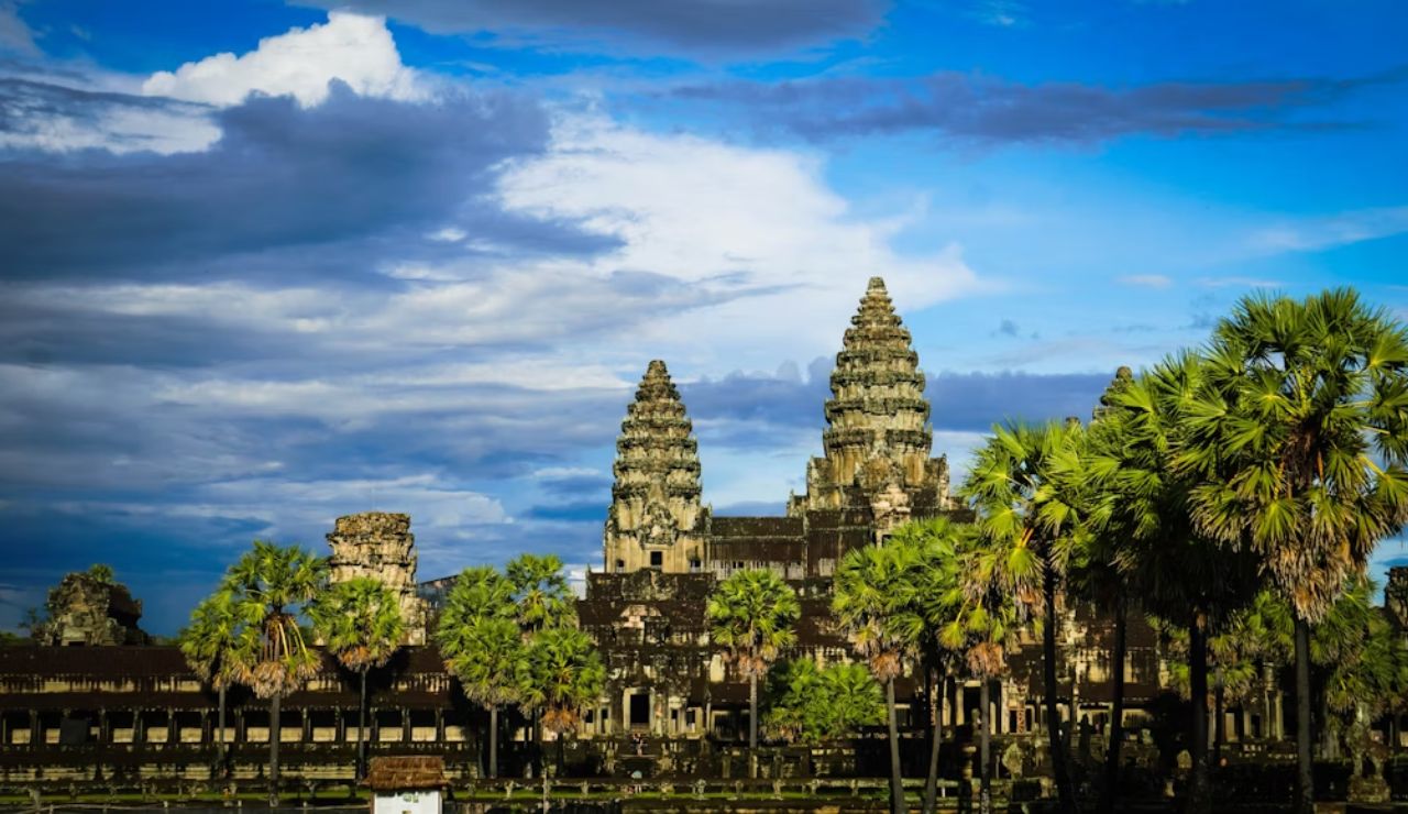 Angkor Wat temple complex in Siem Reap, Cambodia, with palm trees in the foreground under a dramatic blue sky.