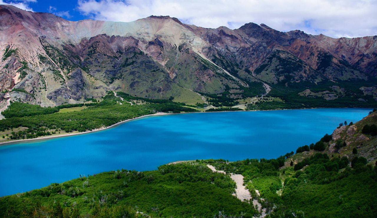 A vibrant blue lake surrounded by lush greenery and rugged mountains under a partly cloudy sky.