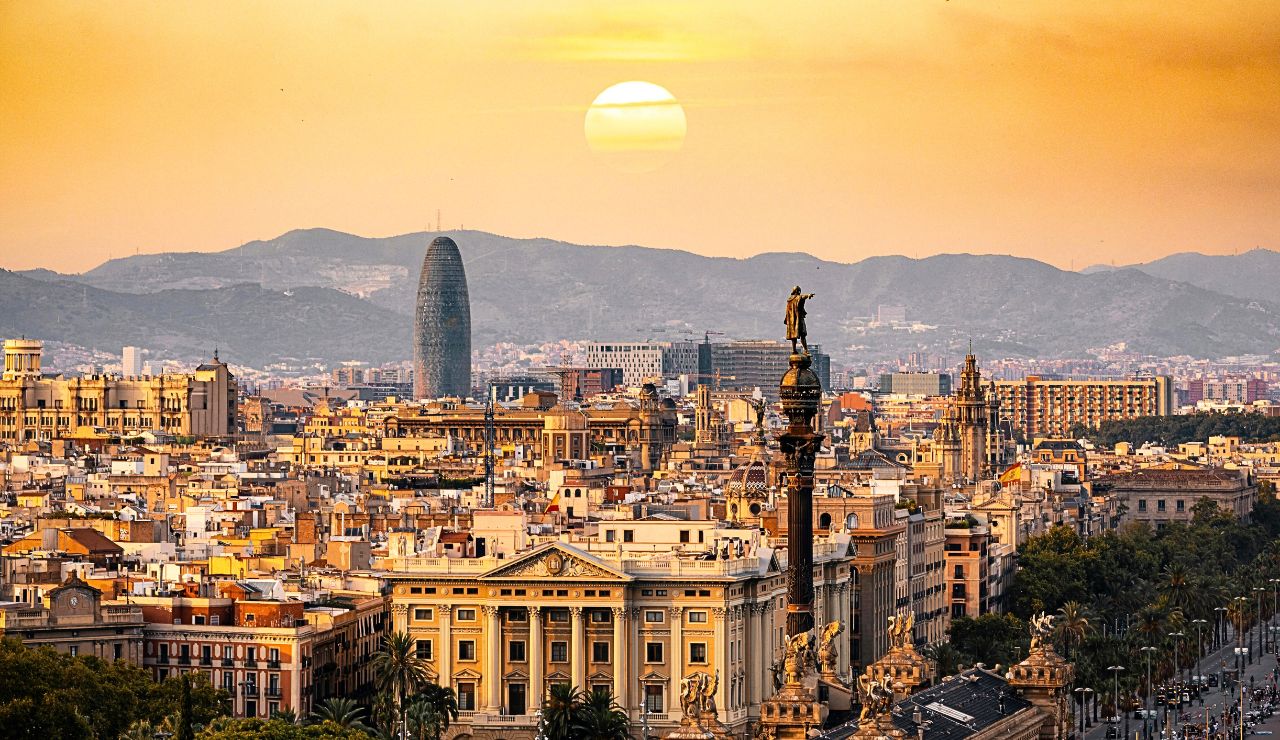A panoramic sunset view of Barcelona shows city buildings, the Columbus Monument, and distant mountains beneath a golden sky.
