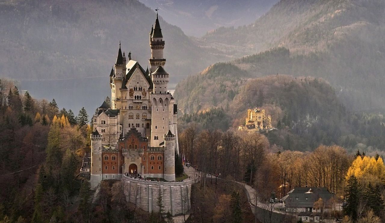 Aerial view of Neuschwanstein Castle in Bavaria, Germany, surrounded by forested hills and mountains with Hohenschwangau Castle visible in the background.