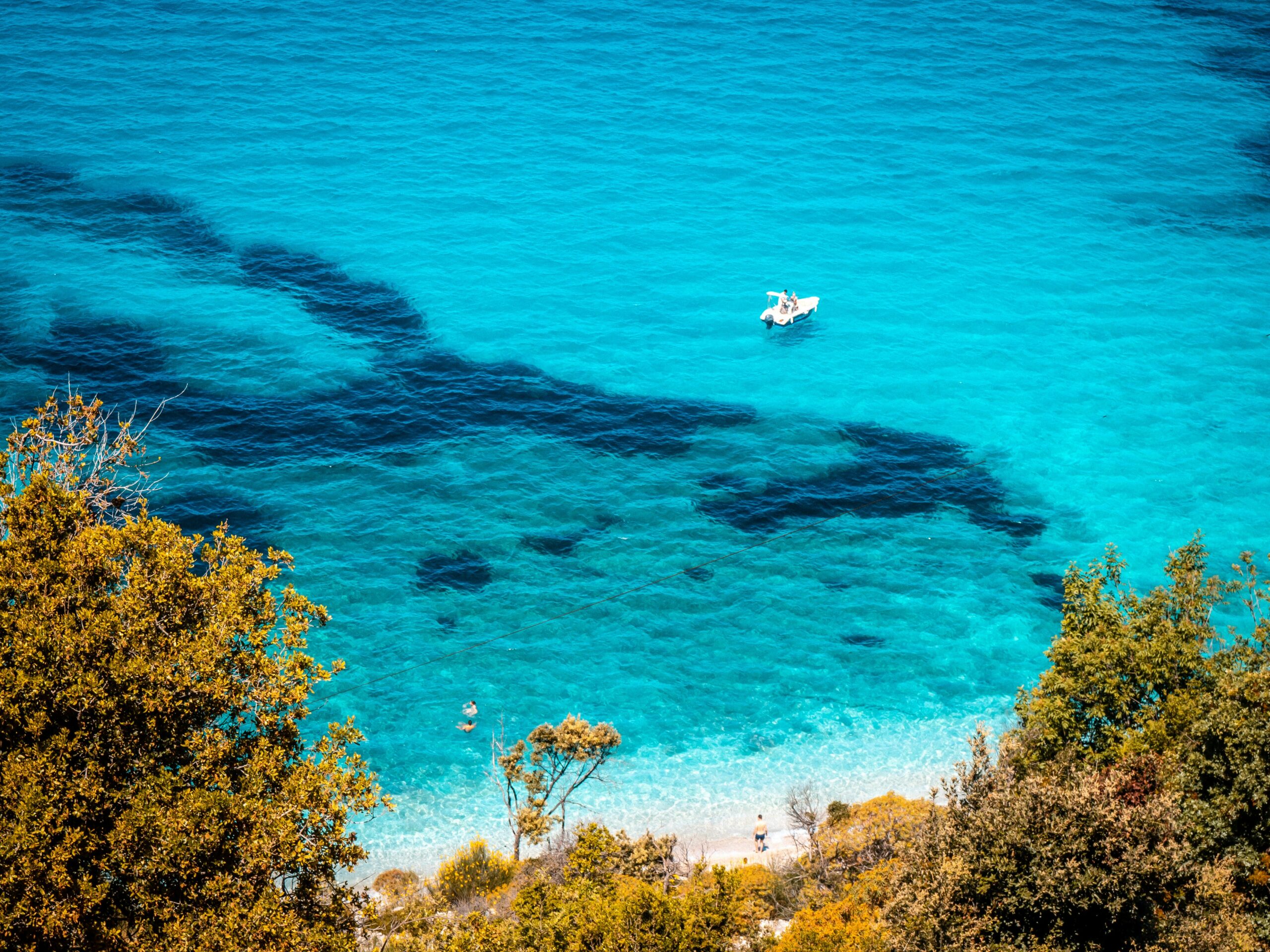 A small boat floats on clear turquoise water near a pebbly shore framed by trees and shrubs.