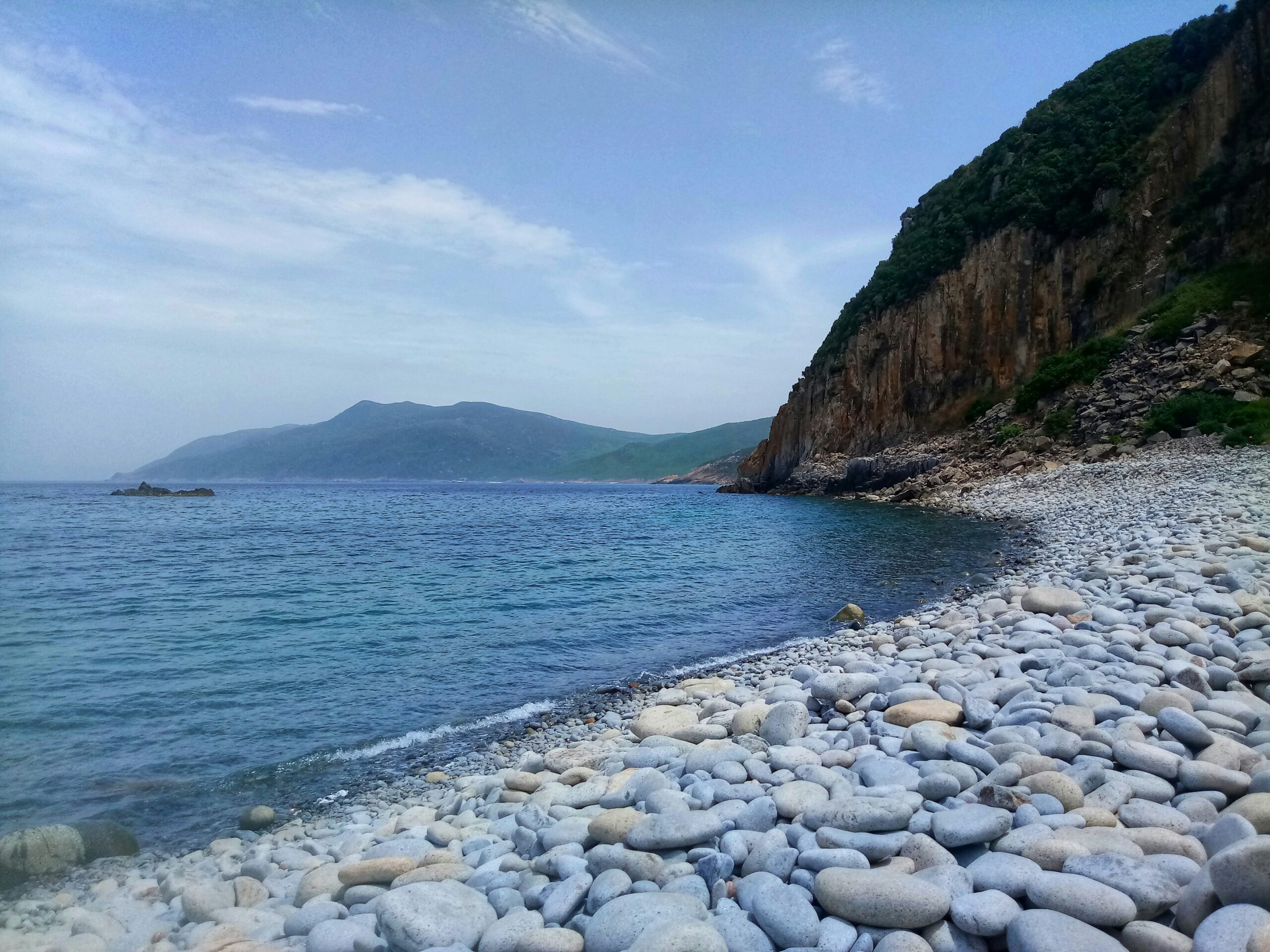 Smooth gray stones on a rocky shore meet clear blue water below a steep, green cliff and partly cloudy sky.