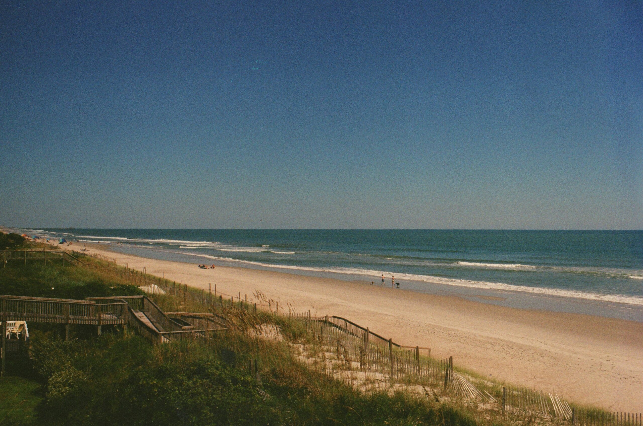 Wooden boardwalks lead to a sandy beach where a few people walk along the shore under a clear blue sky.