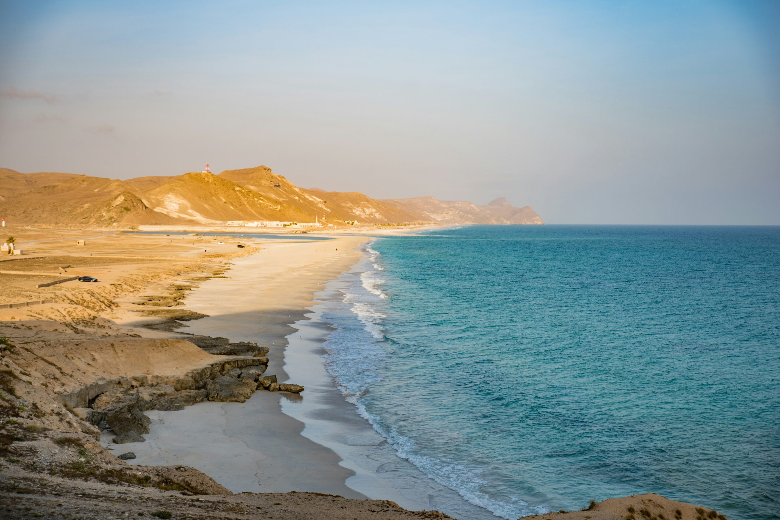 Gentle waves lap a sandy beach bordered by rocky cliffs and distant hills beneath a clear sky.