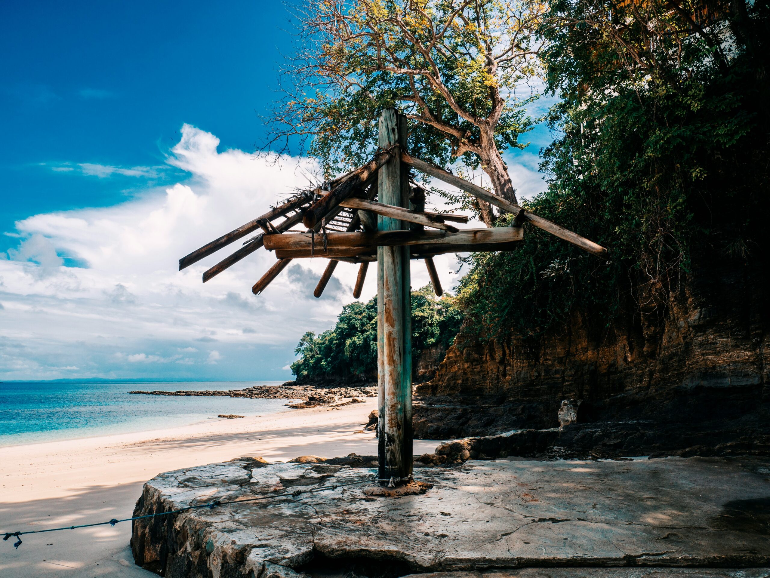 A weathered wooden structure stands on rocks by a sandy beach, with clear blue water, a tree, and a partly cloudy sky in the background.