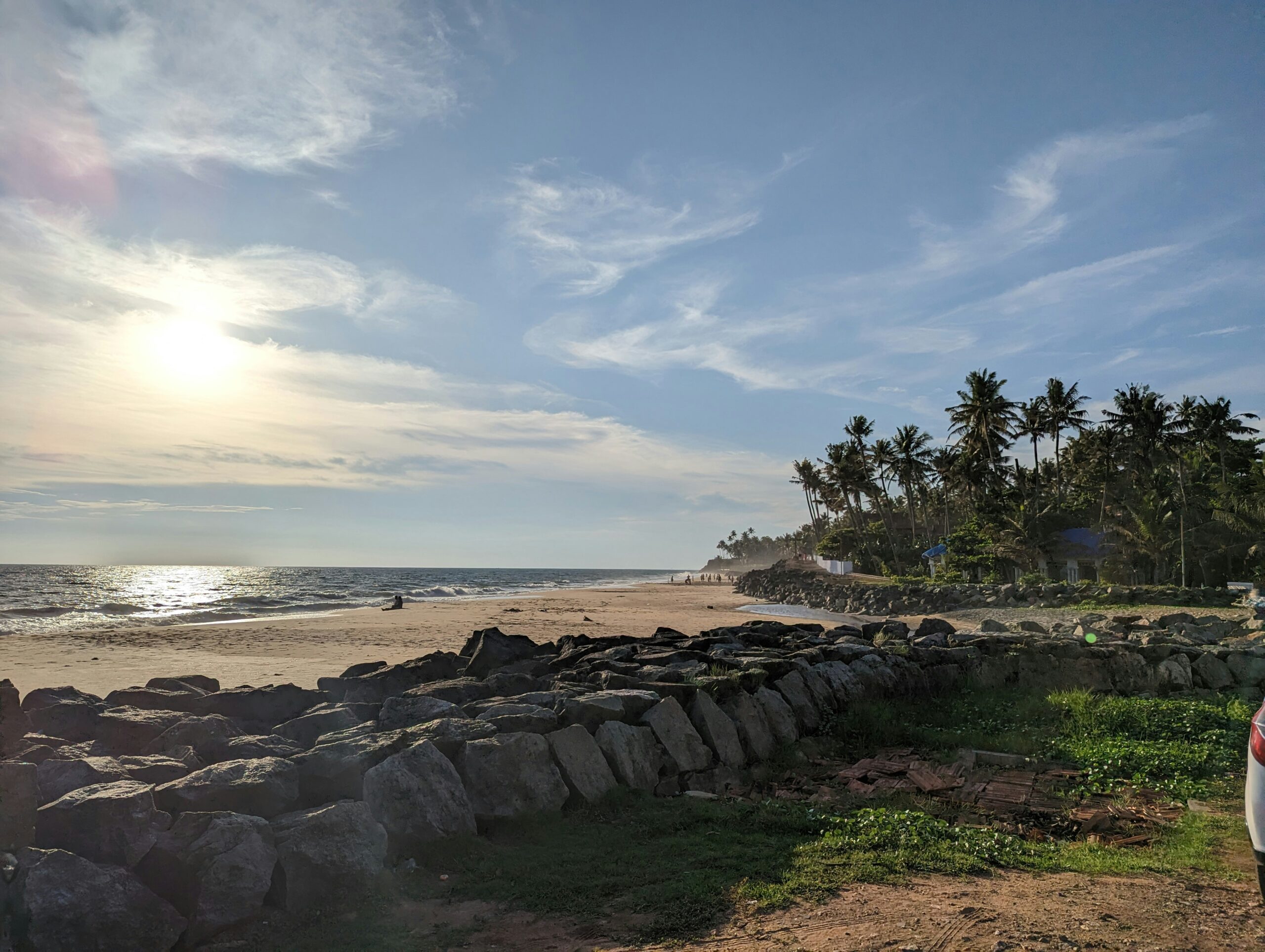 A rocky coastline with palm trees and a sandy beach is illuminated by the low sun over the ocean.