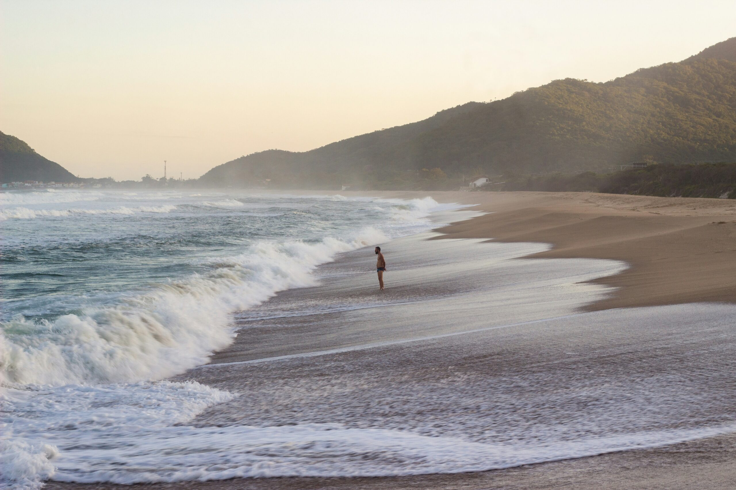 A solitary figure stands at the shoreline of a sandy beach, hills and mist looming in the gentle early light.