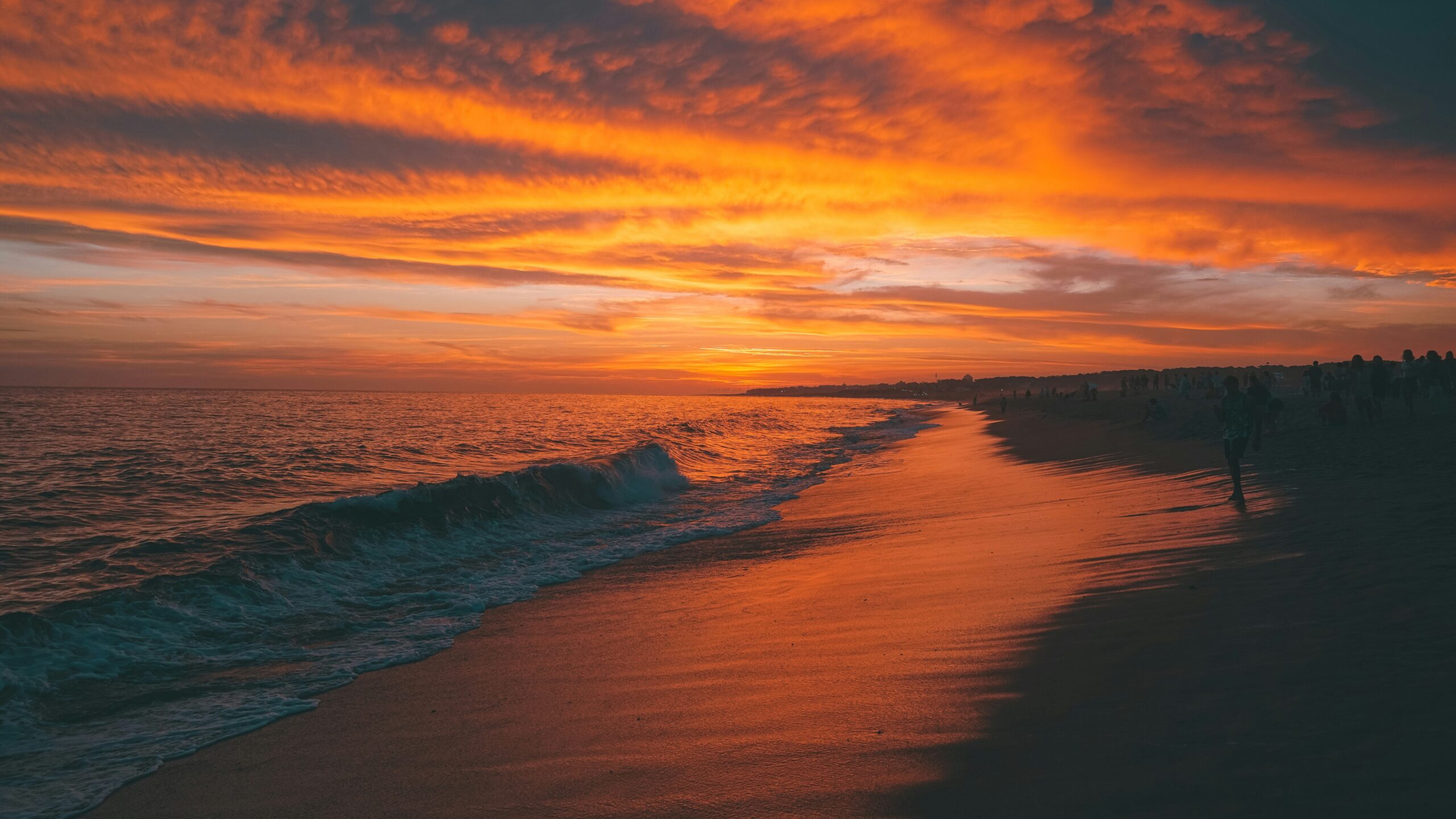 A vivid orange-red sunset illuminates ocean waves washing onto a sandy beach, silhouetting distant people.