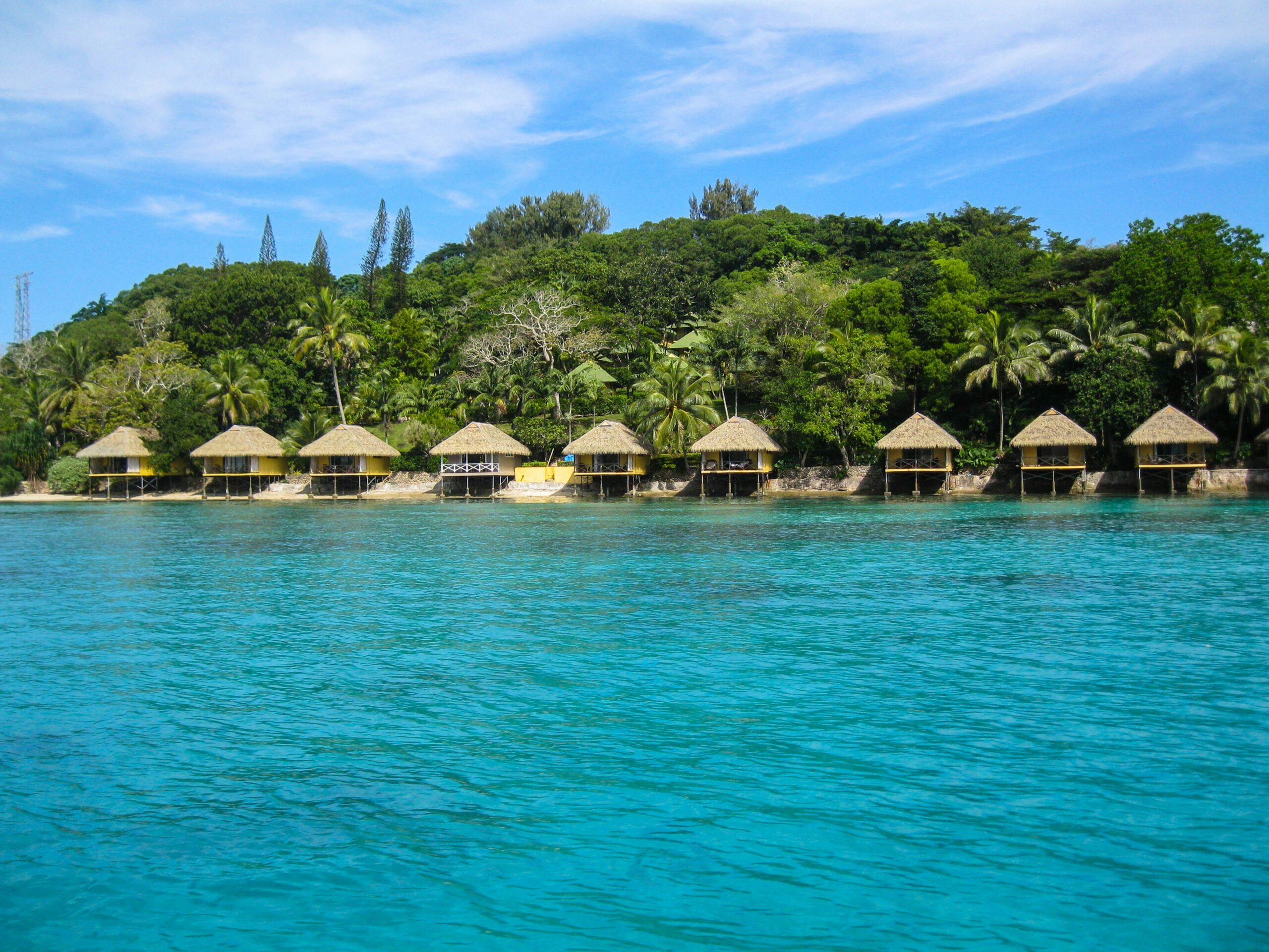 Overwater thatched-roof bungalows line a lush shoreline above clear turquoise water beneath a partly cloudy sky.