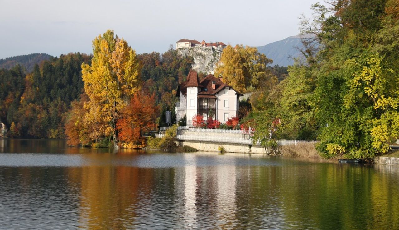 Scenic view of Lake Bled in Slovenia with a lakeside villa surrounded by colorful autumn trees and Bled Castle perched on a cliff in the background.