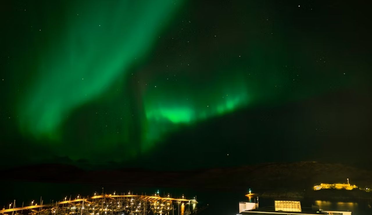 Northern lights glowing over a marina and coastal buildings in Bodø, Norway.