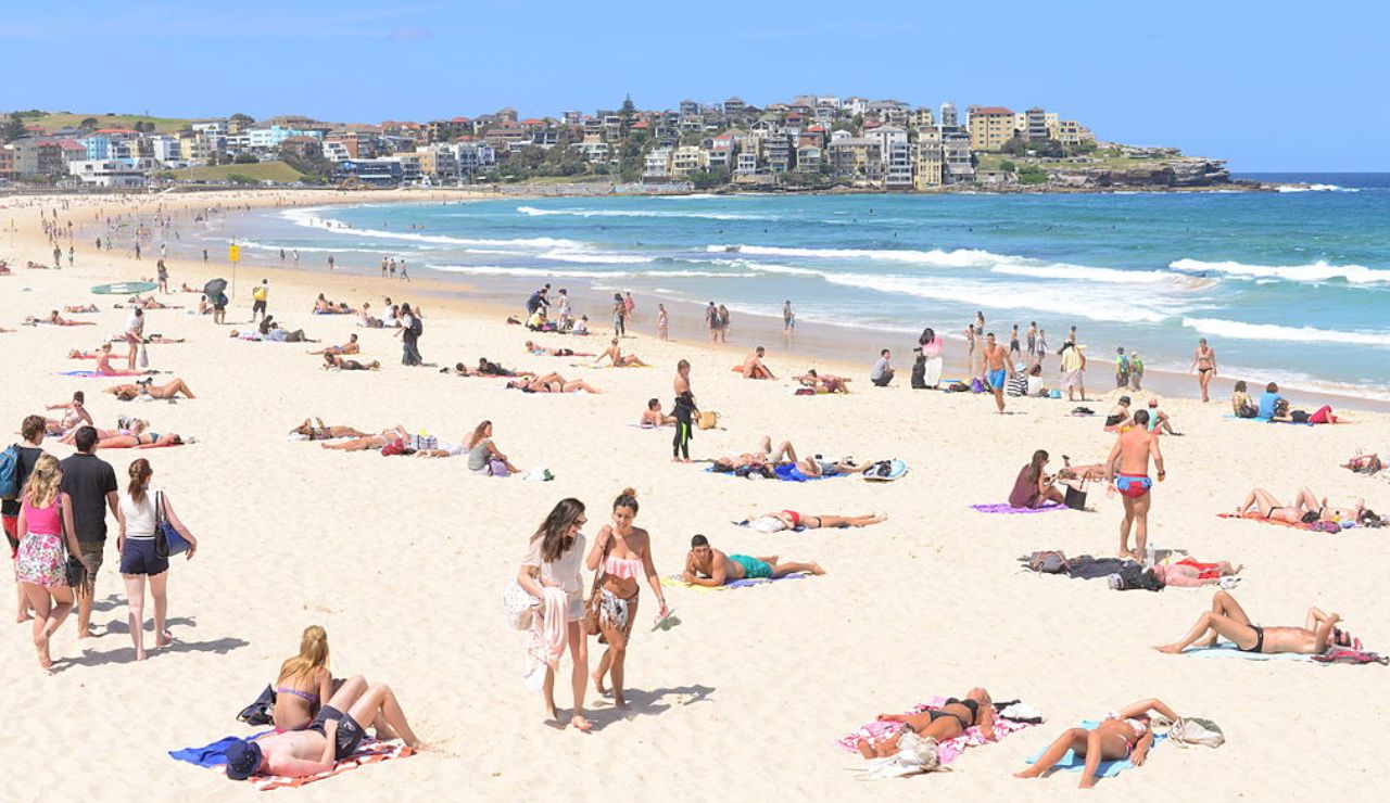 Beachgoers sunbathe and stroll along the sandy shore of Bondi Beach on a bright, sunny day.