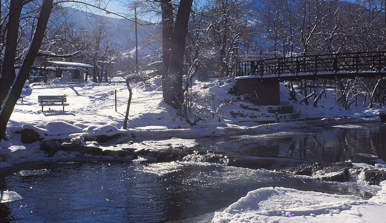 Snow-covered park with a wooden footbridge over a partially frozen creek and mountains in the background on a sunny winter day