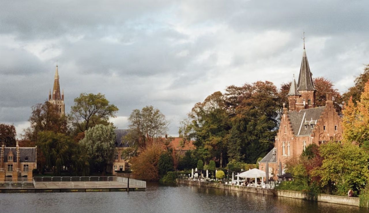 Scenic view of Minnewater Castle and surrounding buildings along the canal in Bruges, Belgium, with autumn trees and a cloudy sky in the background.