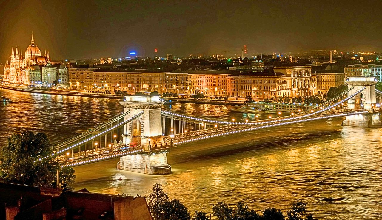 Night view of the illuminated Chain Bridge over the Danube in Budapest, with city buildings and the Hungarian Parliament in the background.