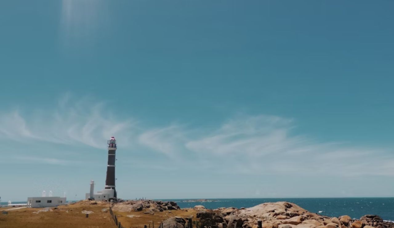 Rustic lighthouse standing on the rocky coastline of Cabo Polonio, Uruguay, under a clear blue sky with ocean waves in the distance.