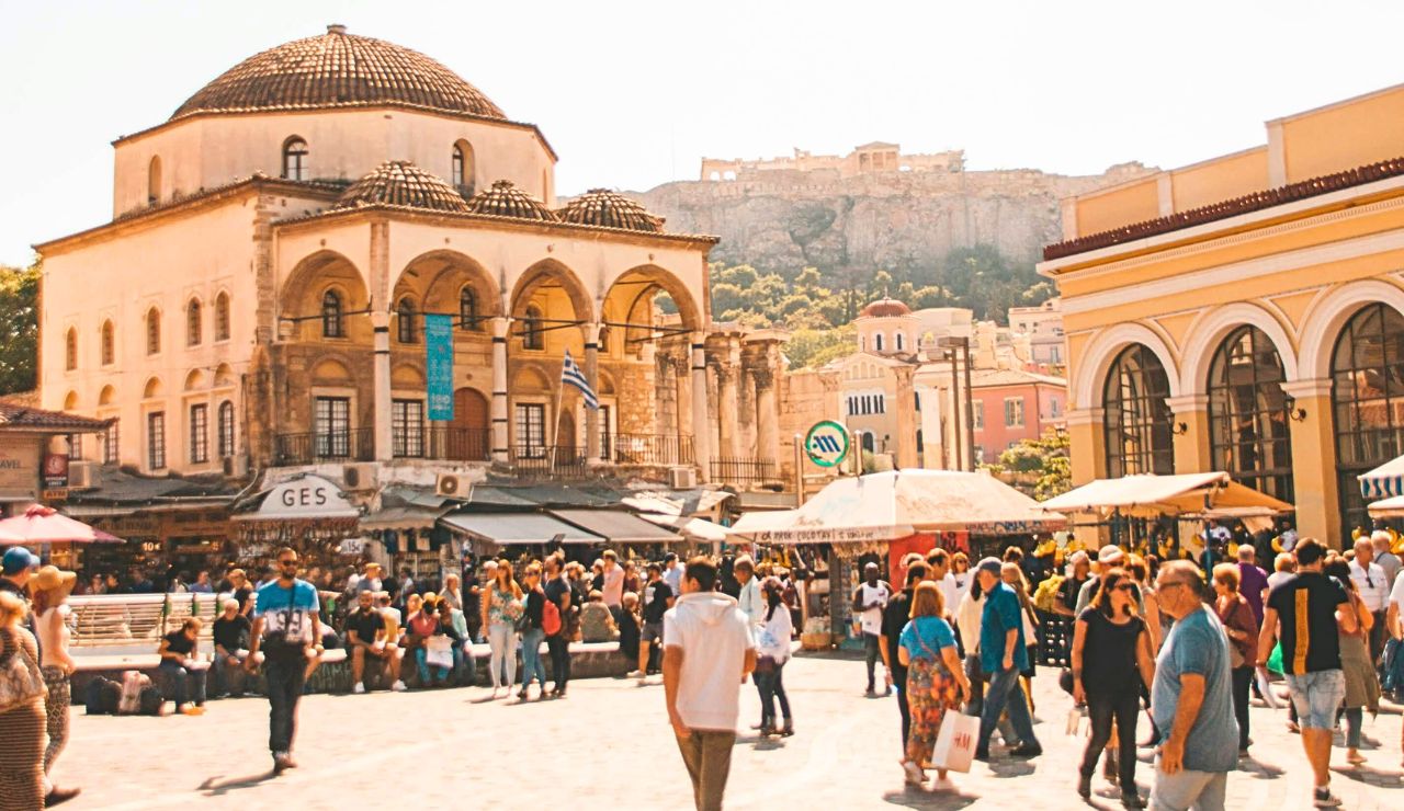 Crowds fill a busy square lined with historic buildings and a domed mosque, with the Acropolis visible on the hill behind.