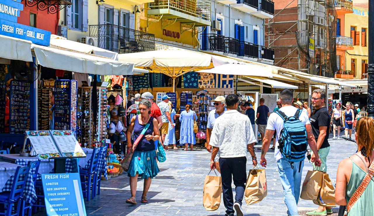 People stroll through a bustling outdoor market street lined with shops and restaurants, browsing or carrying shopping bags beneath sun umbrellas.