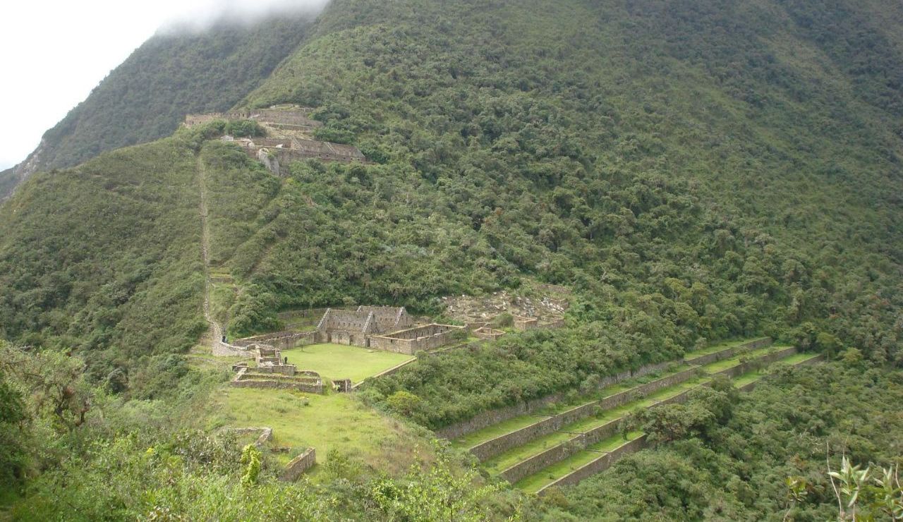 Ancient stone ruins lie nestled within lush green mountains, with terraced slopes and mist brushing the upper hillsides.