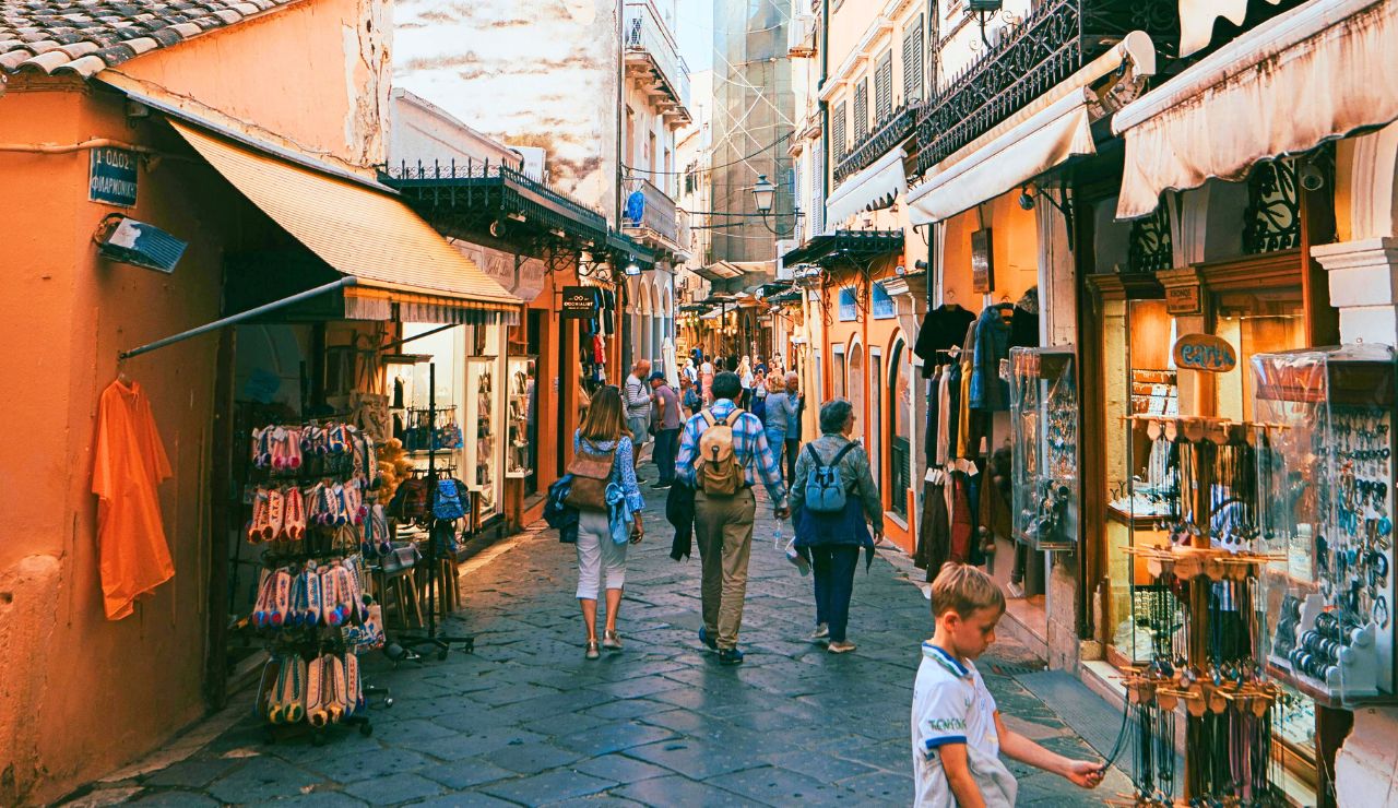 People walk along a narrow cobblestone street lined with shops selling souvenirs, clothes, and jewelry in a busy outdoor market.
