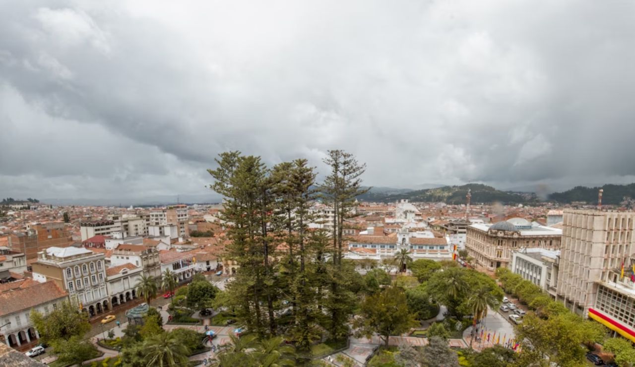 Panoramic view of Cuenca, Ecuador, showcasing the historic center, colonial architecture, and central plaza with tall trees under a cloudy sky.