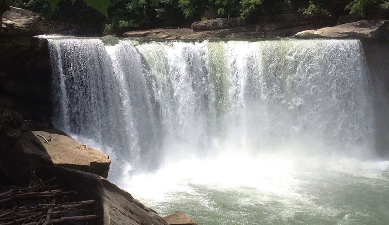 A wide, powerful waterfall plunges over a rock ledge, sending mist into the air as it crashes into the greenish pool below.