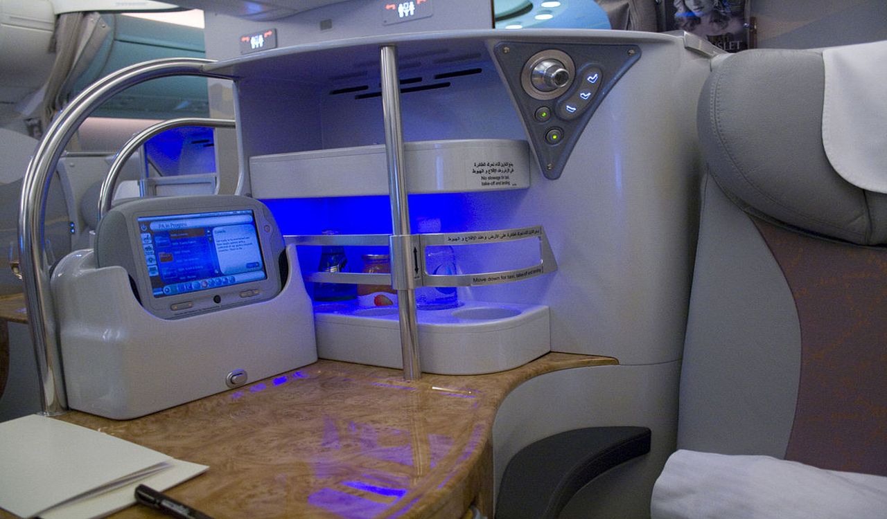 A luxurious airline seat setup featuring a sleek entertainment screen, ambient lighting, personal storage, and a polished wood-finish table—ideal for comfort and productivity in-flight.