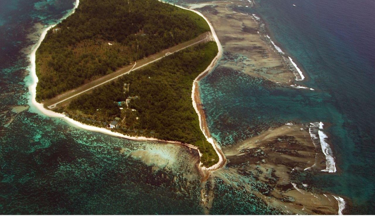 A remote island surrounded by coral reefs, with a narrow airstrip running through dense tropical forest and turquoise waters hugging the shore.