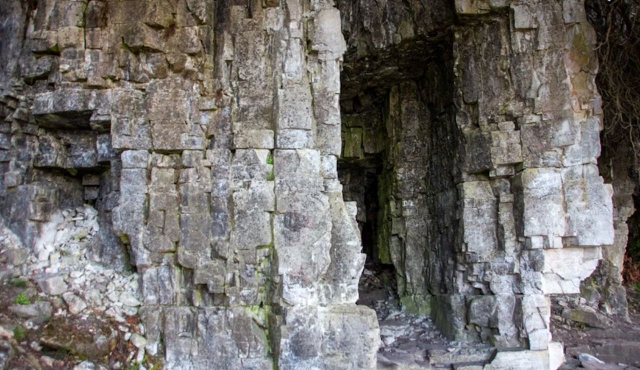 Entrance to a rugged limestone cave with angular rock formations and weathered stone walls, partially covered in moss and surrounded by loose rubble.