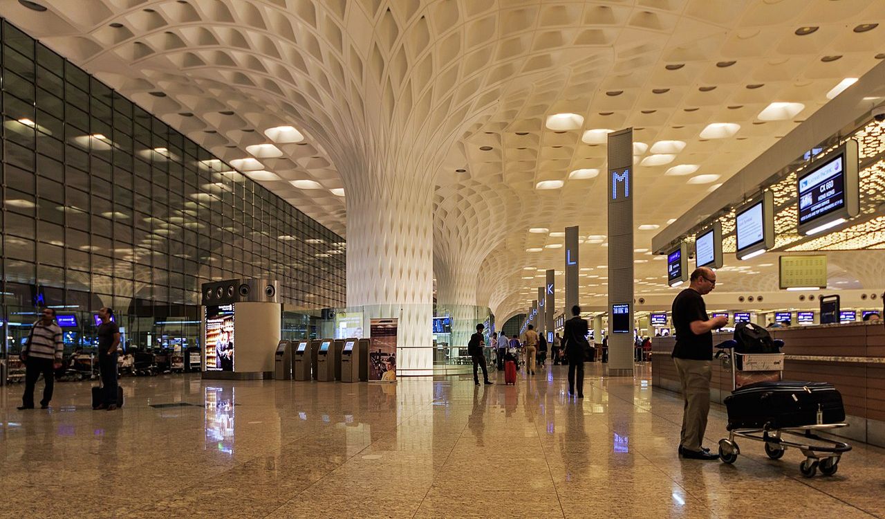A modern airport terminal features stunning architectural columns and a polished floor, with passengers checking in and moving with their luggage.