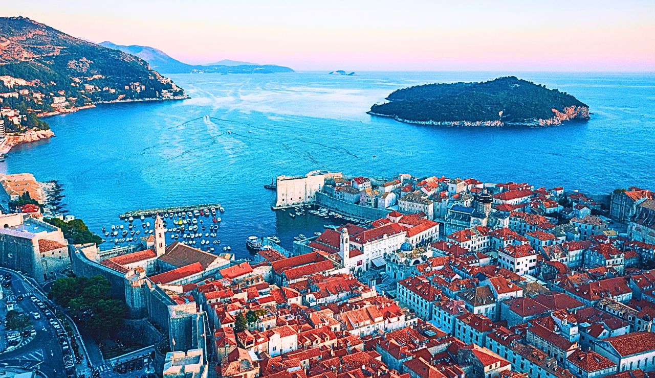 Aerial view of Dubrovnik, Croatia, features red-roofed buildings, historic city walls, and the blue Adriatic Sea with nearby islands at sunset.