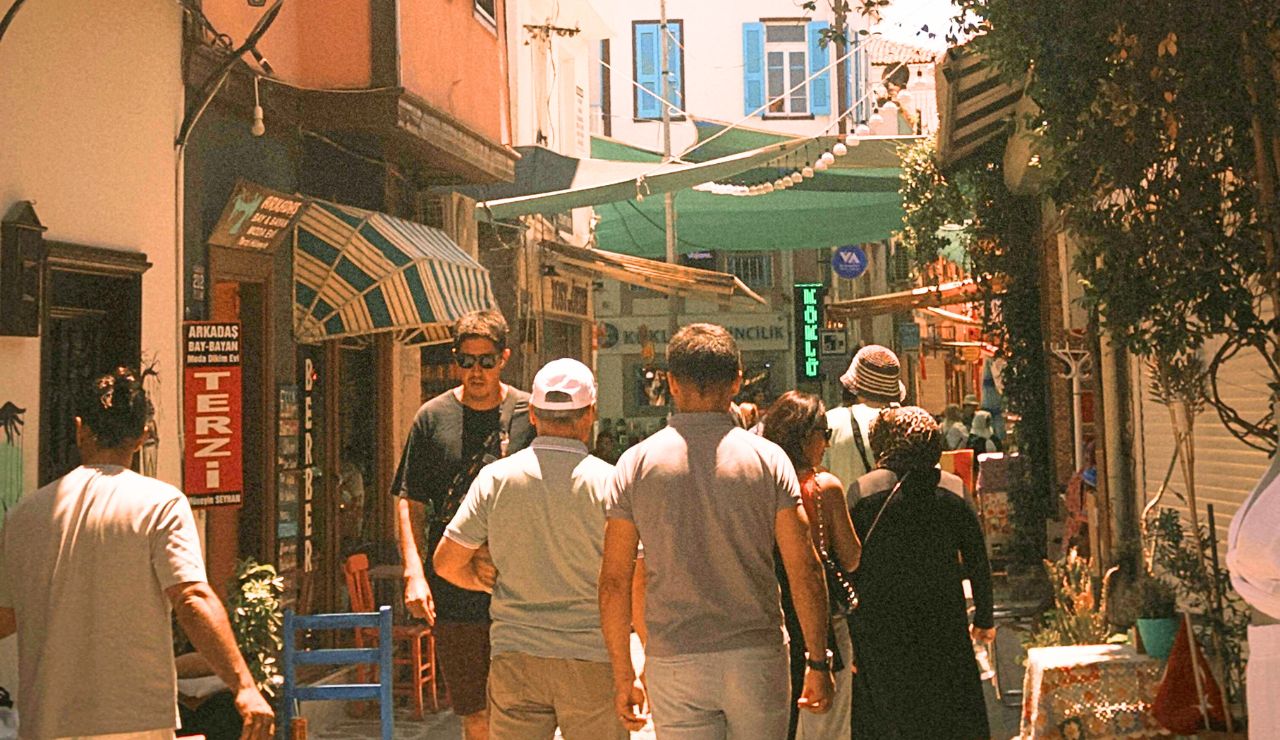 People walk down a narrow, sunlit street lined with shops, colorful signs, and window-shuttered buildings.
