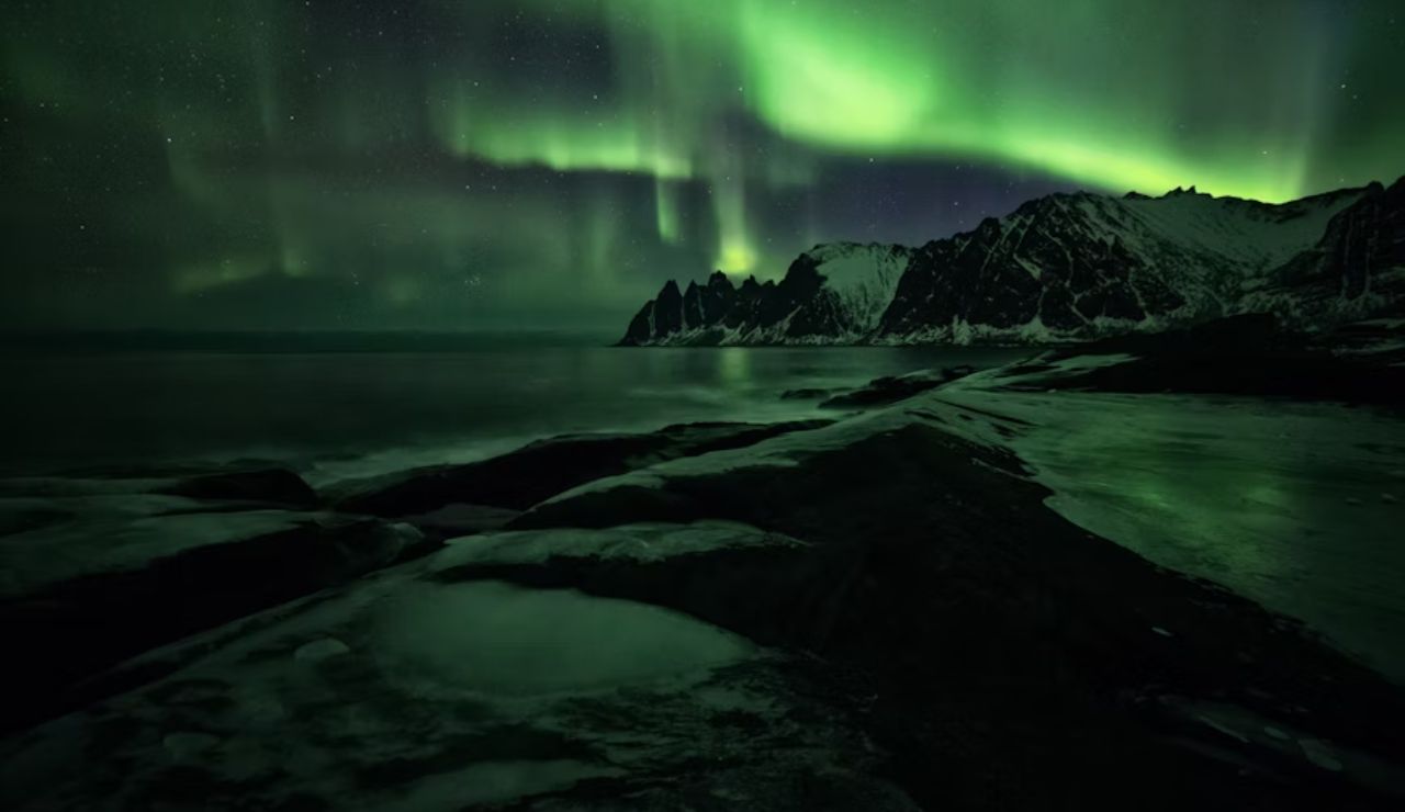 Northern lights glowing above snow-covered mountains and icy shoreline in Ersfjordbotn, Norway.