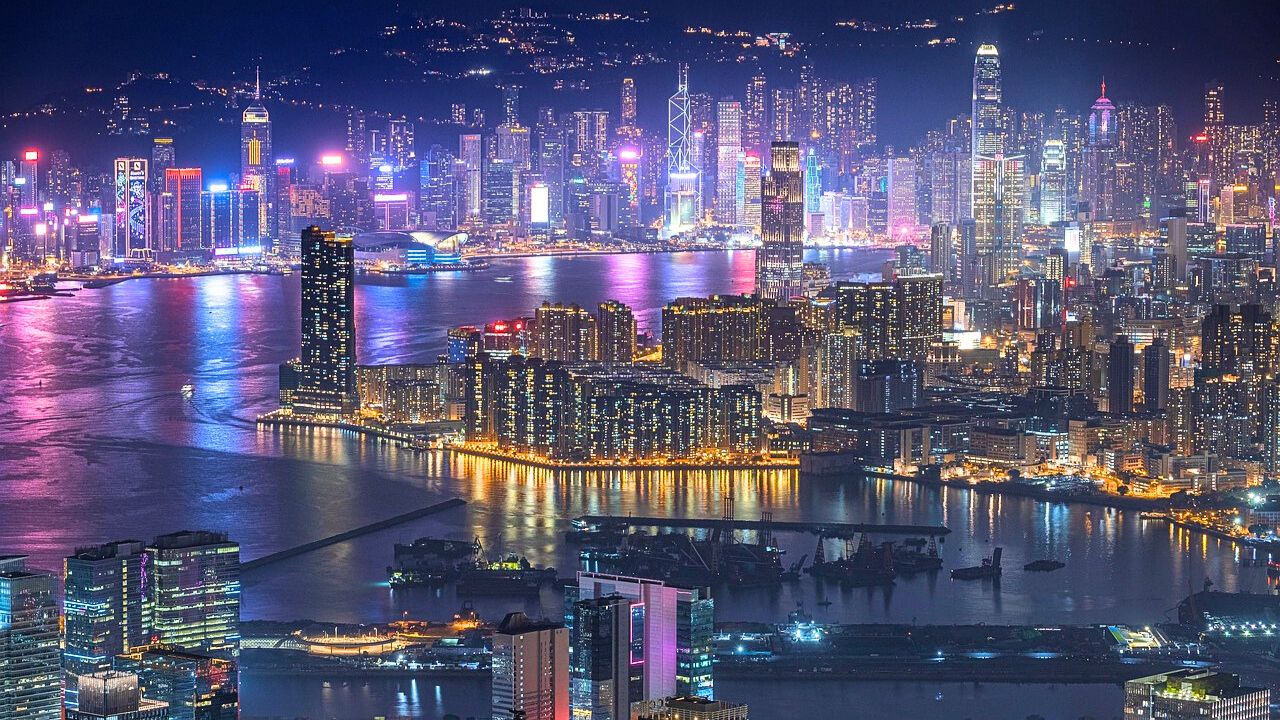 Aerial night view of a densely built Hong Kong skyline with brightly lit skyscrapers reflecting on the water.