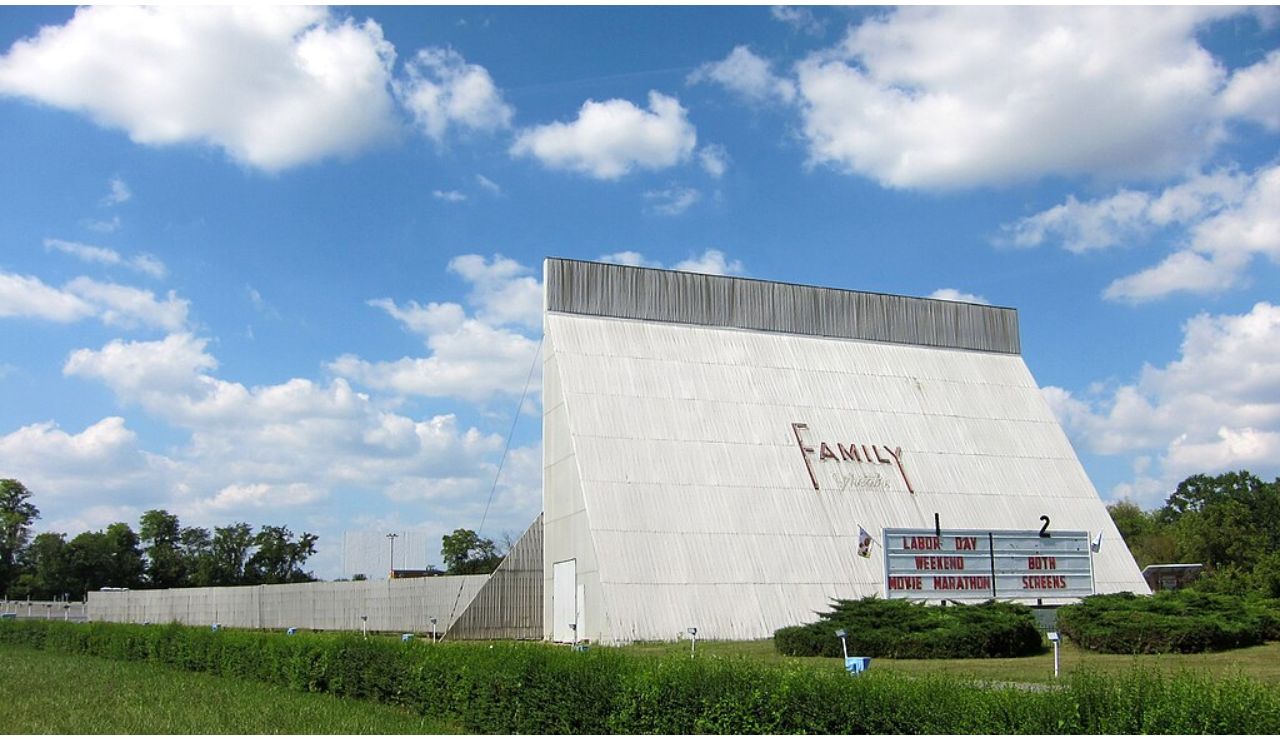 Family Drive-In Theatre with a large white screen and marquee promoting a Labor Day weekend movie marathon on both screens, set under a bright blue sky.