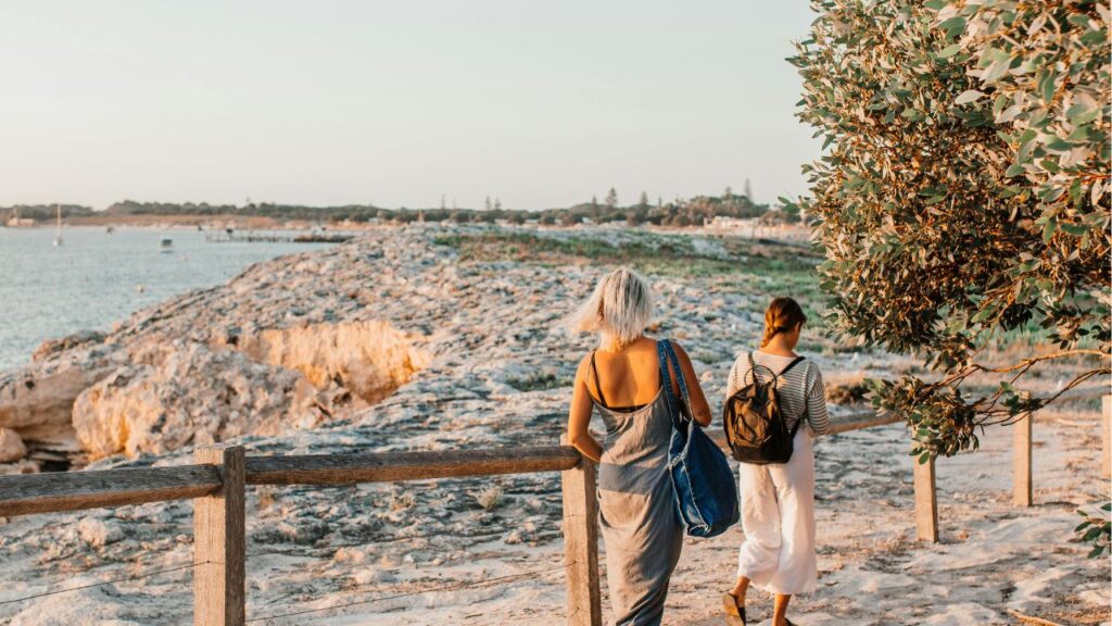 Two people walk along a rocky coastal path bordered by trees and a wooden fence near the ocean.