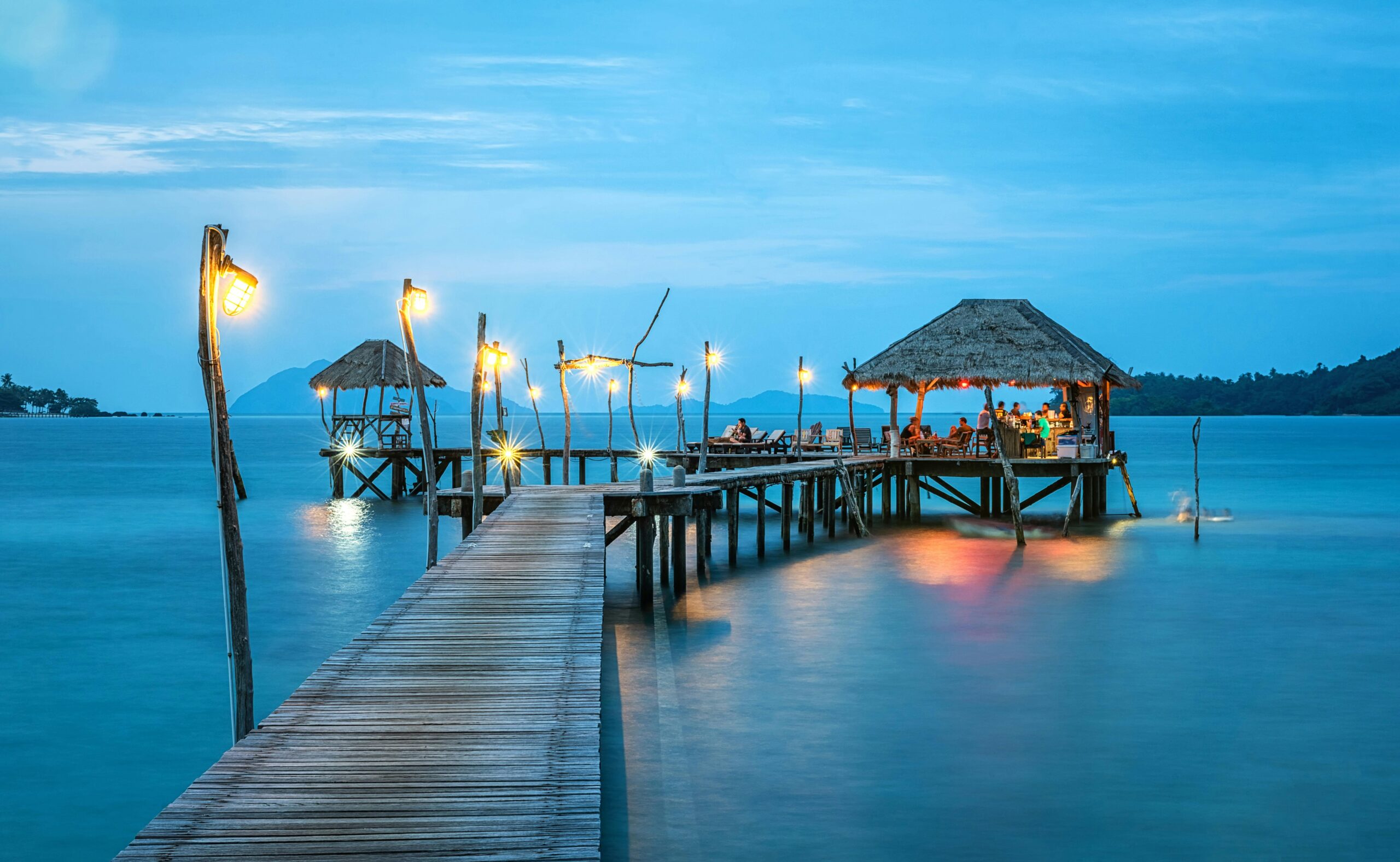 At dusk, a wooden pier with thatched-roof huts and illuminated lights extends over calm blue water, where people sit at tables.