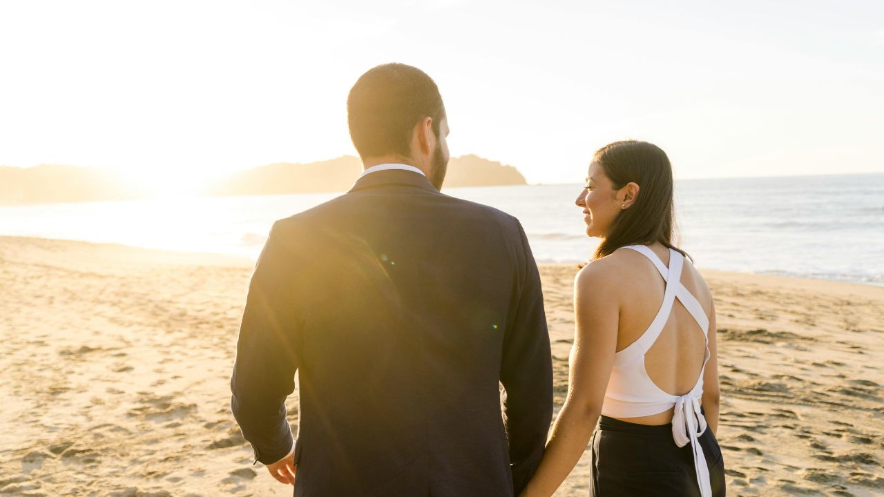 A man and woman walk hand in hand along a sandy beach at sunset, facing the ocean.
