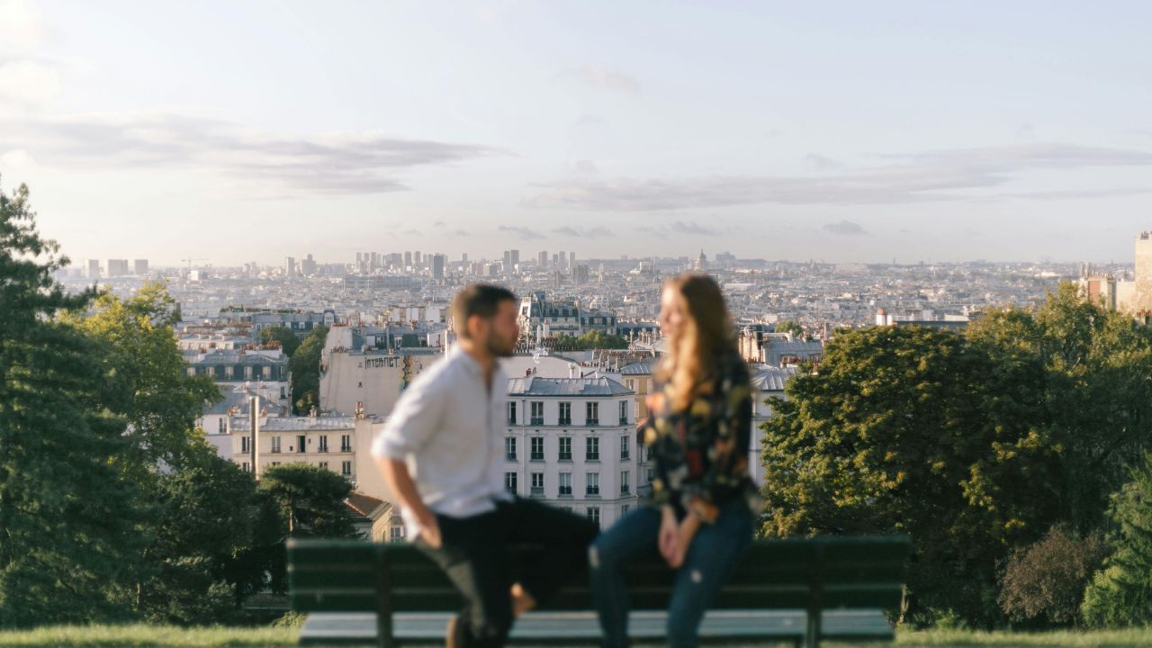 Two people sit on a bench in the foreground with a blurred city skyline behind them under a partly cloudy sky.