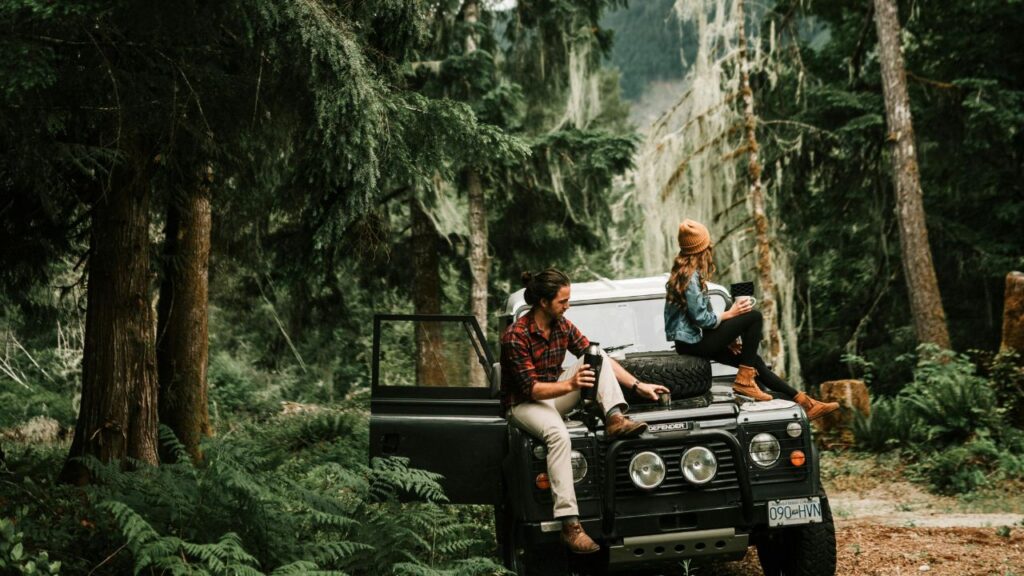 Two people sit on the hood of a parked off-road vehicle in a forest, holding cups and looking in different directions.