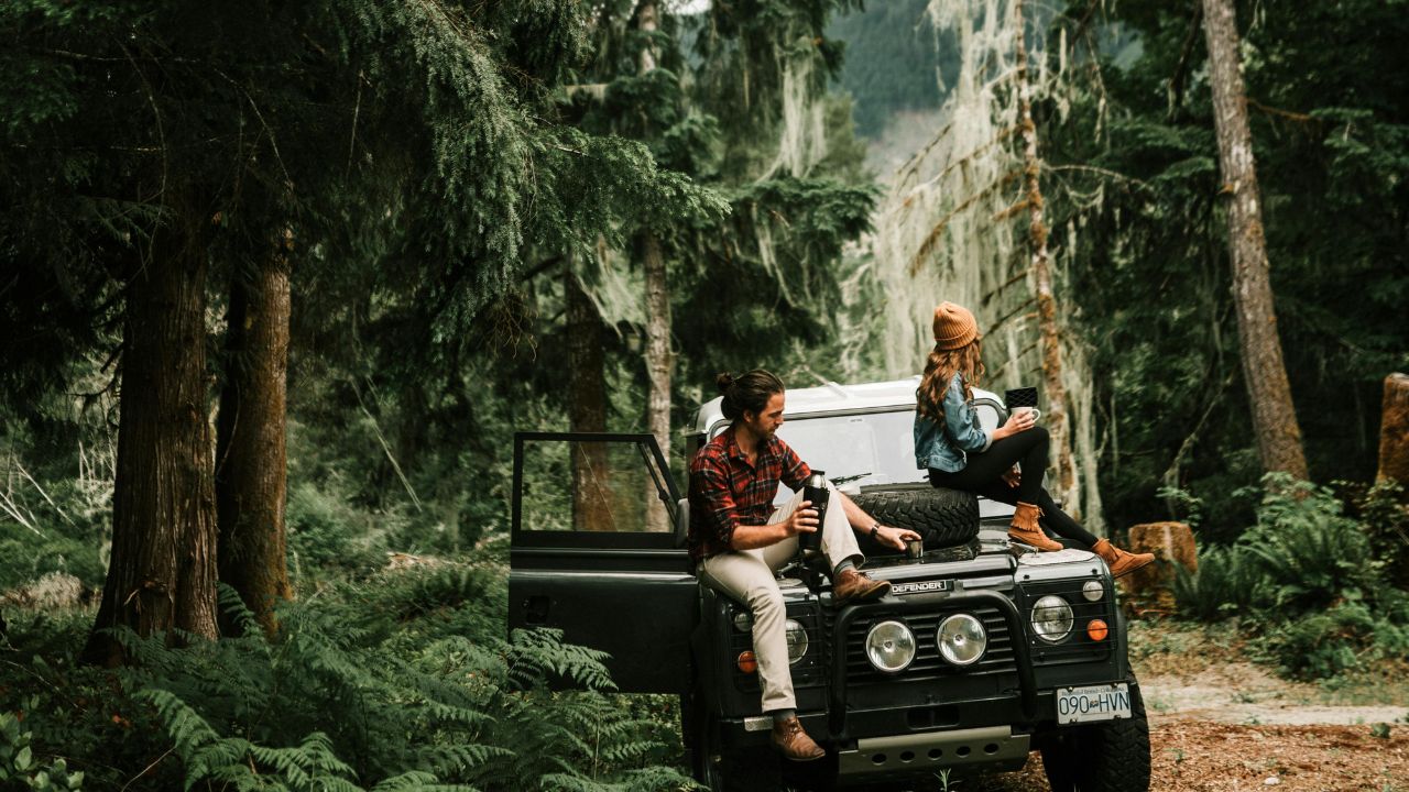 Two people sit on the hood of a parked off-road vehicle in a forest, holding cups and looking in different directions.