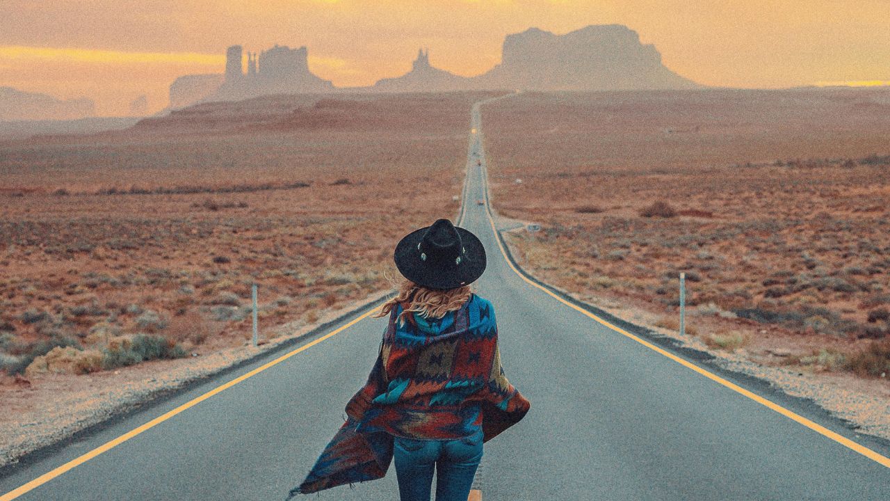 A person in a hat and patterned poncho stands on an empty desert road at sunset, facing distant rock formations.